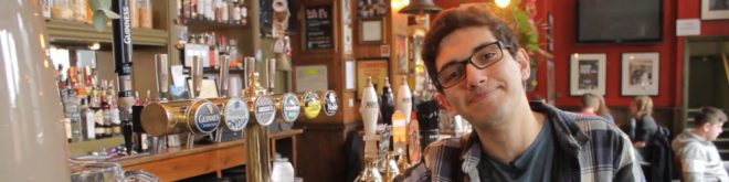 Homem em pub tradicional de Londres ao lado de chopeiras com várias opções de cervejas, incluindo Guinness.