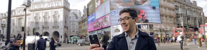 Homem sorridente tirando selfie em Piccadilly Circus, Londres, com telões coloridos e ônibus vermelhos ao fundo.