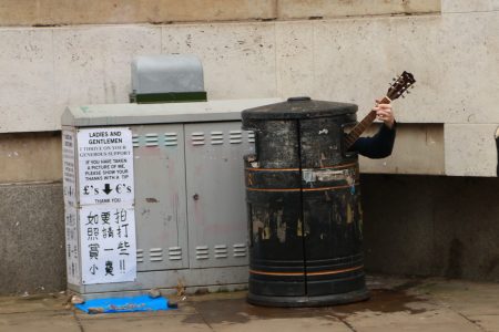 artista de rua tocando dentro de lata de lixo em cambridge com uma placa em vários idiomas pedindo uma gorejta