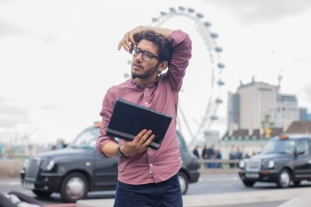 Homem com camisa rosa e óculos segura uma pasta enquanto olha pensativo, com a London Eye e táxis pretos de Londres ao fundo.