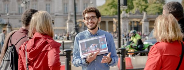 Guia turístico sorridente mostra foto histórica a visitantes em frente ao Palácio de Buckingham, em Londres.
