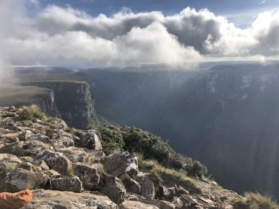 Vista panorâmica de cânion cercado por montanhas verdes, com nuvens baixas e raios de sol atravessando o céu.