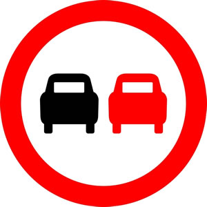 Placa de trânsito com dois carros lado a lado, um preto e outro vermelho, dentro de um círculo vermelho, indicando “proibido ultrapassar”.