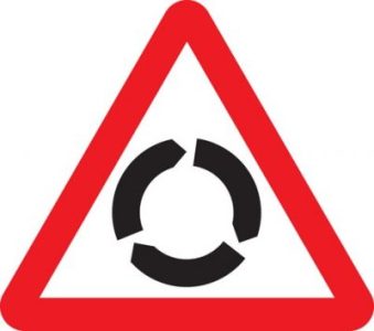 Placa de trânsito triangular com borda vermelha e símbolo preto circular formado por setas, indicando “rotatória à frente” ou “aproximação de interseção em sentido circular.