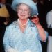 rainha mãe elizabeth bebendo uma taça de vinho com uma roupa azul e levantando a taça como quem está brindando.