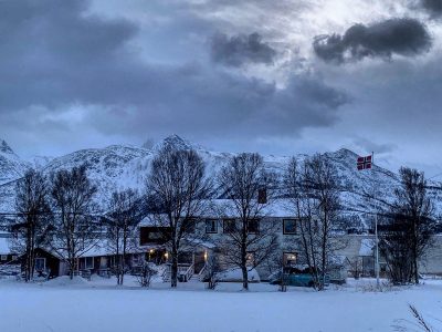 casas com neve em cima, árvores sem folhas, montanhas com neve e a bandeira da noruega