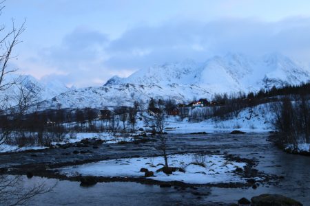 montanhas com neve e um pequeno vilarejo com luzes acesas e um rio em frente, no norte da noruega