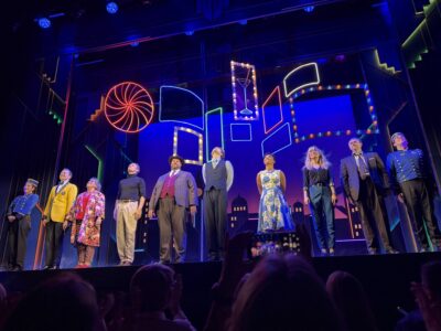 Elenco de peça de teatro com roupas de época da década de 40 e funcionários de hotel num palco com iluminação azul no West End de Londres agradecendo o público da noite