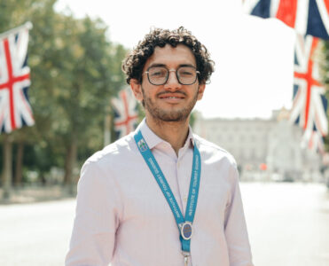 Guia de turismo Rafael Maciel com credencial de guia de turismo em Londres no pescoço. Credencial azul clara de guia Blue Badge, com bandeiras britânicas e palácio de Buckingham ao fundo
